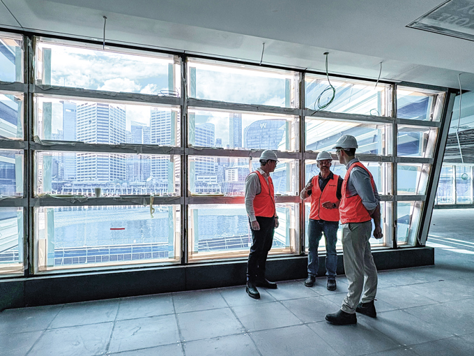 Three construction workers in orange vests and helmets discuss inside a building under construction with large windows overlooking a city skyline.