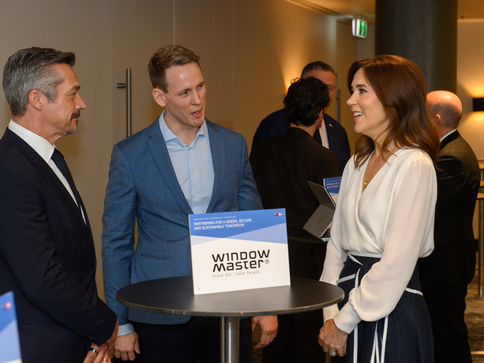People in business attire stand and converse around a high table with a "WindowMaster" sign in a conference or networking event.