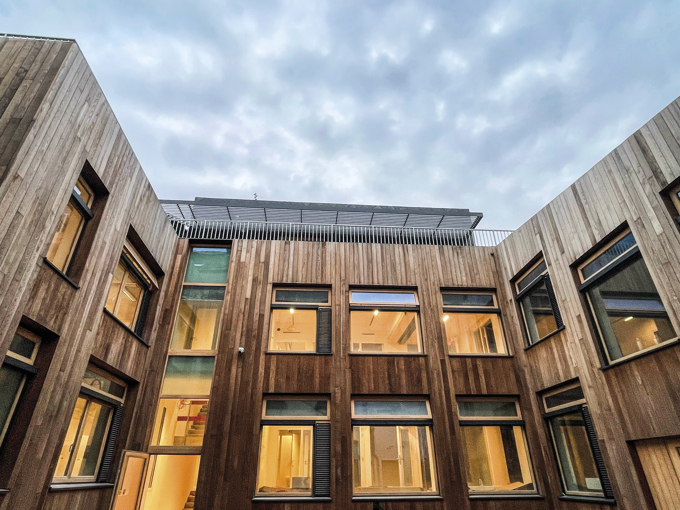 Modern wooden building courtyard with large windows under a cloudy sky at dusk, interior lights glowing warmly.