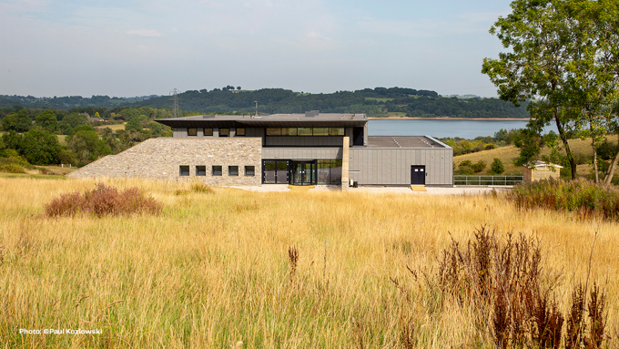 Modern building with stone and glass facade, surrounded by tall grass and trees, near a lake and green hills under a blue sky.
