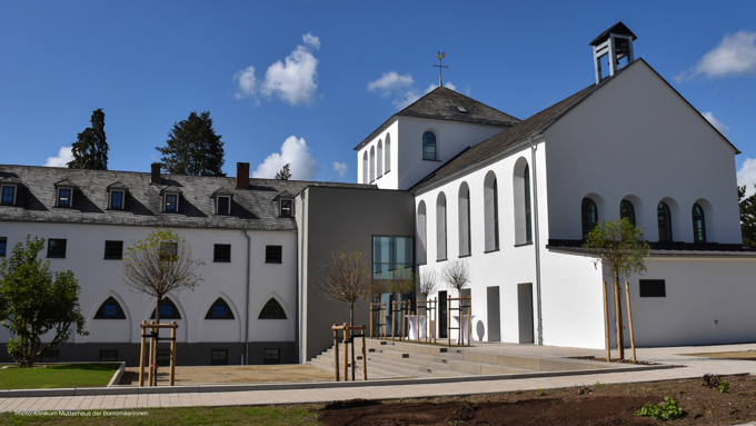 Modern white building featuring arched windows, a slate roof, and landscaped grounds under a blue sky with fluffy clouds.