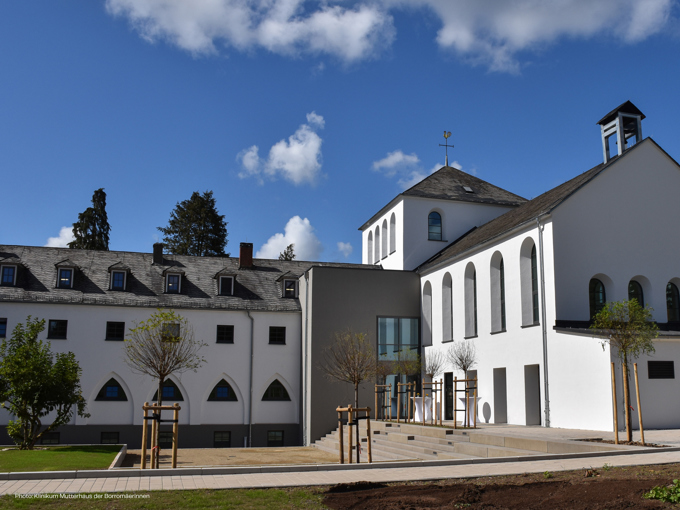 Modern white building featuring arched windows, a slate roof, and landscaped grounds under a blue sky with fluffy clouds.