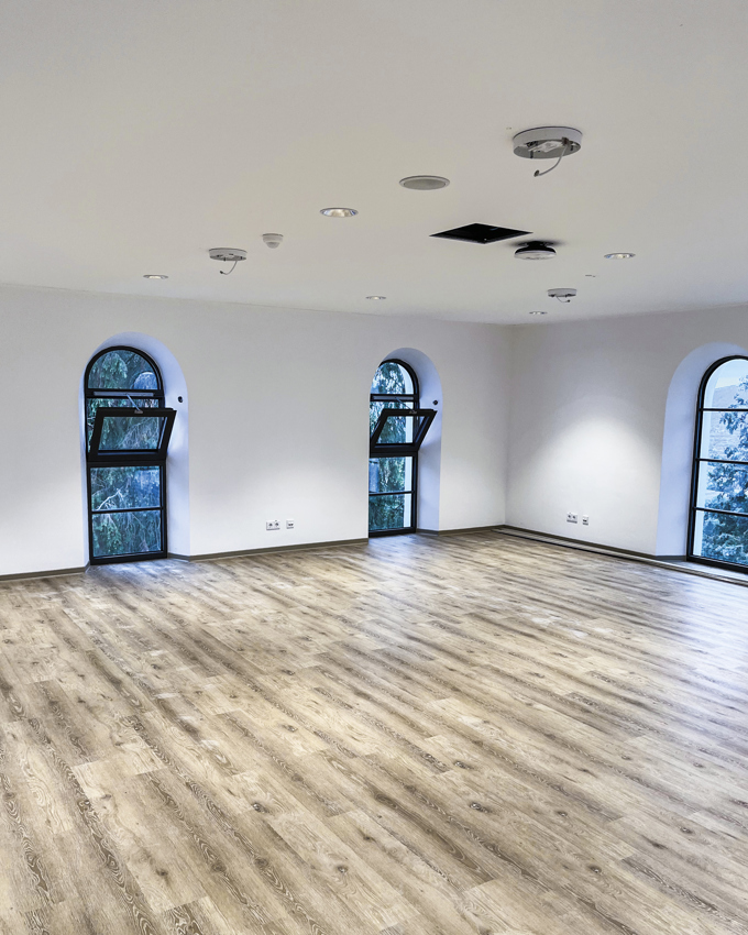 Bright, empty room with wooden flooring and arched windows, featuring white walls and modern ceiling lights.