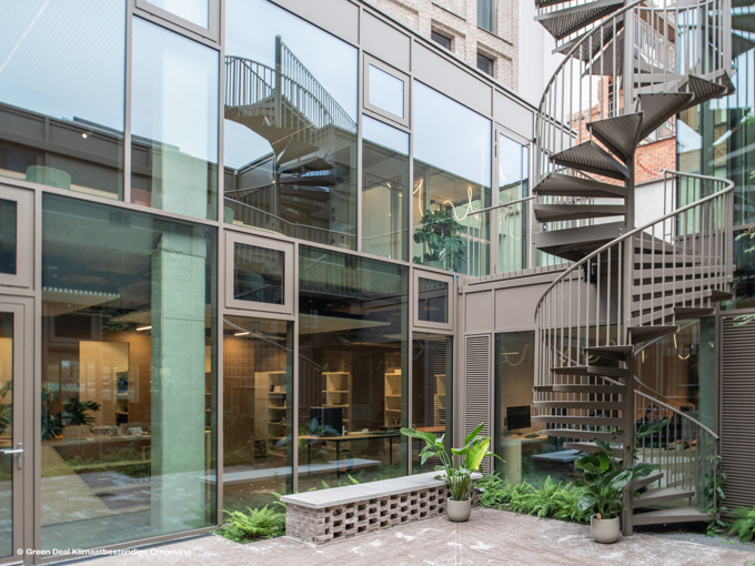 Contemporary outdoor courtyard featuring modern glass buildings, a spiral staircase, and lush greenery in planters.