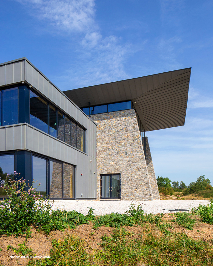 Modern building featuring a stone facade and large glass windows, topped with an angular metal roof against a blue sky.