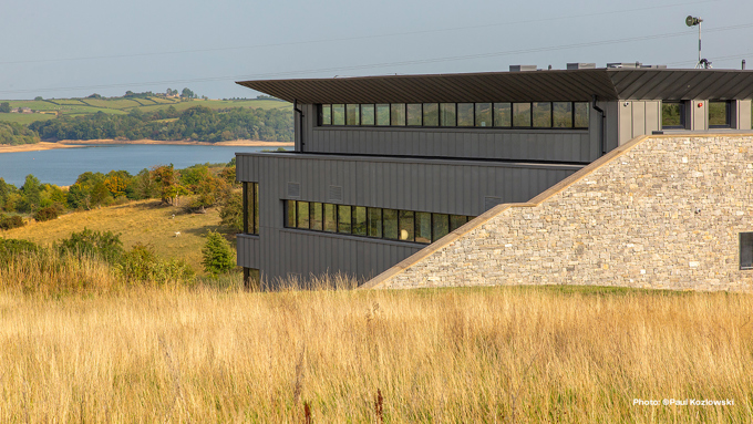 Modern building with a flat roof and large windows, overlooking a river and lush green hills on a clear day.