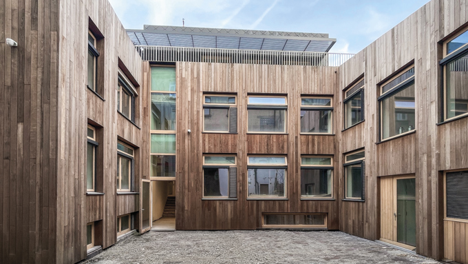 Courtyard surrounded by modern wooden-paneled buildings with multiple windows and a paved ground under a cloudy sky.