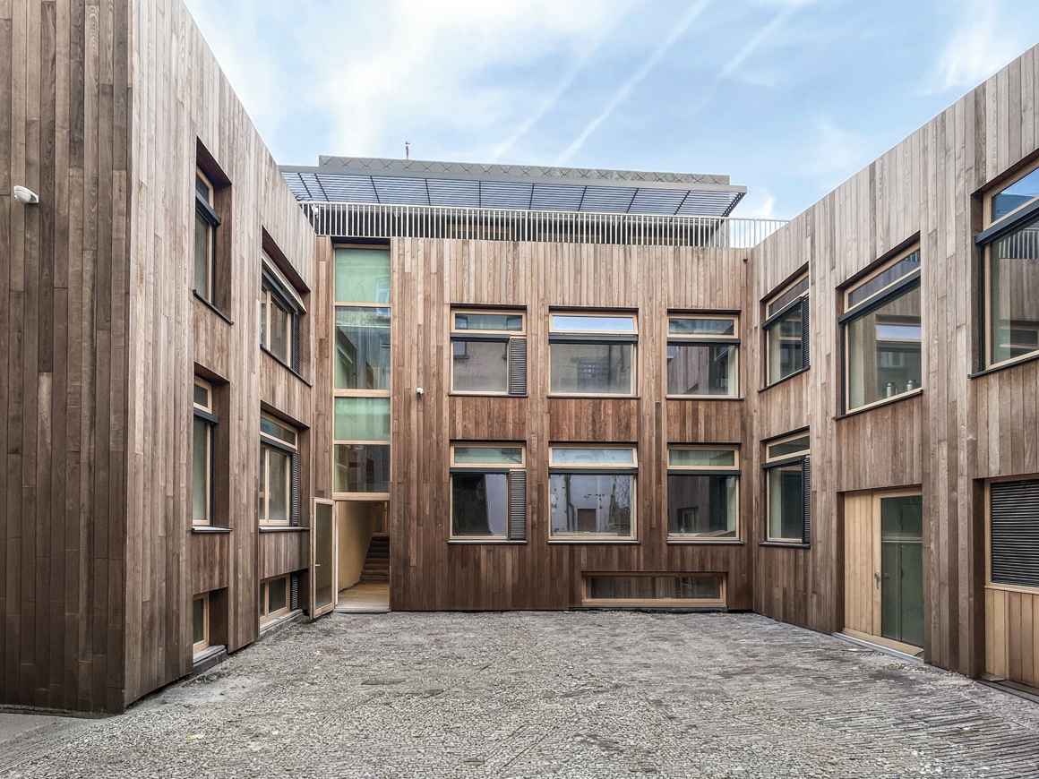 Courtyard surrounded by modern wooden-paneled buildings with multiple windows and a paved ground under a cloudy sky.