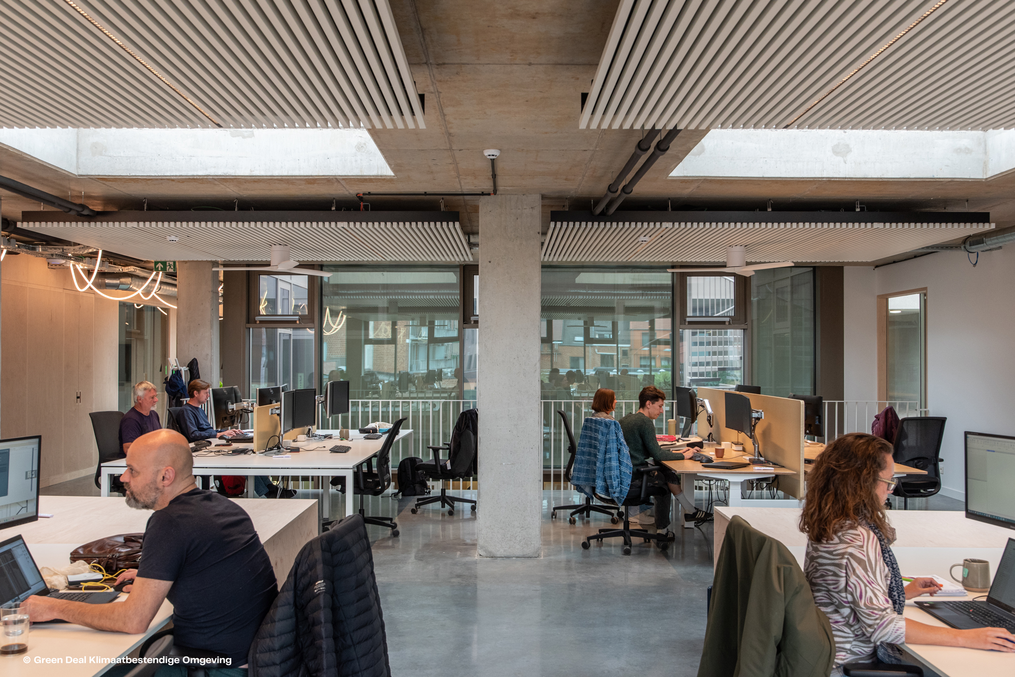 A modern office space featuring several desks with computers, natural light, and a concrete ceiling, occupied by focused individuals.