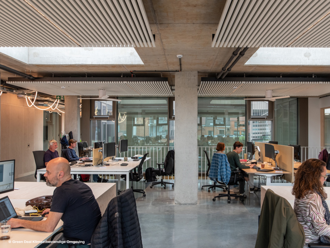 A modern office space featuring several desks with computers, natural light, and a concrete ceiling, occupied by focused individuals.