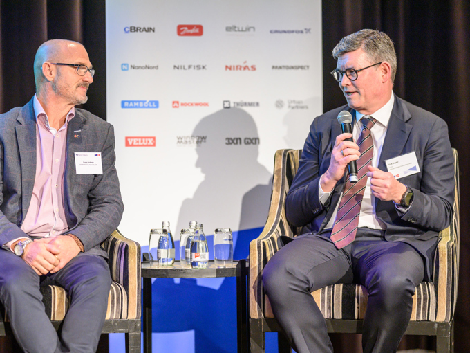 Two men in suits having a discussion on stage, with a microphone, water bottles, and a sponsor board in the background.