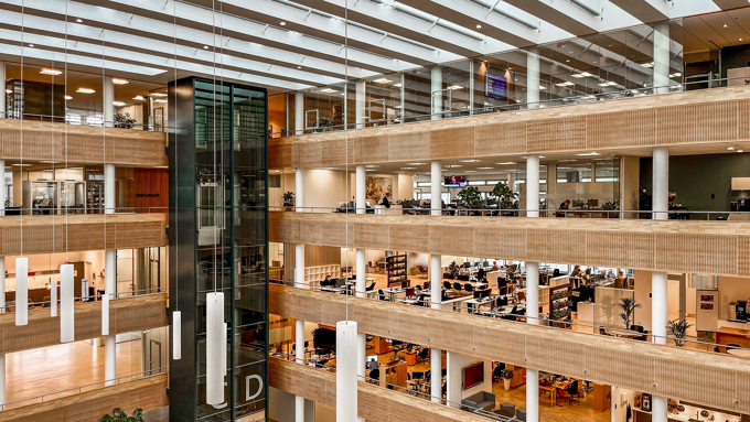Modern office interior featuring multiple floors, an elevator, and large open spaces filled with greenery and workstations.