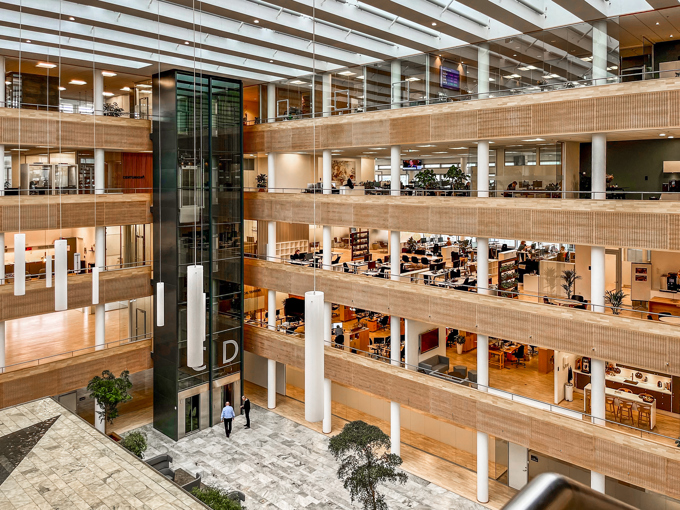 Modern office interior featuring multiple floors, an elevator, and large open spaces filled with greenery and workstations.