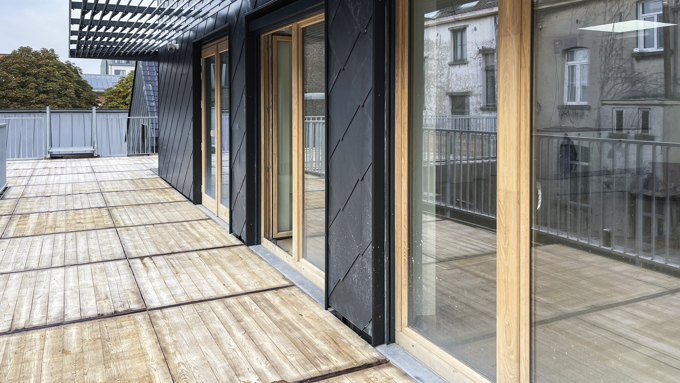 Spacious rooftop terrace with wooden flooring, black-framed glass doors, metal pergola, and a view of neighboring buildings.