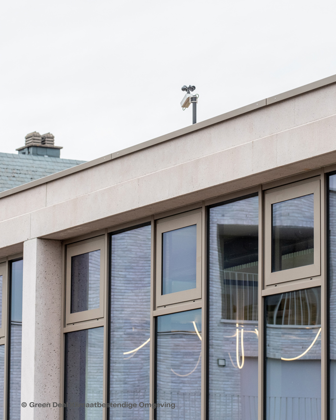 A modern building facade with large windows, showcasing a sleek design and a weather station on the roof against a cloudy sky.
