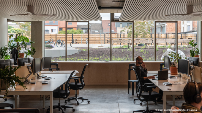A modern office space with desks, computers, and plants, featuring large windows that offer a view of an outdoor garden and bicycles.