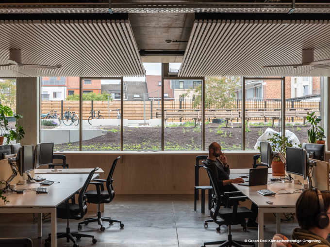 A modern office space with desks, computers, and plants, featuring large windows that offer a view of an outdoor garden and bicycles.