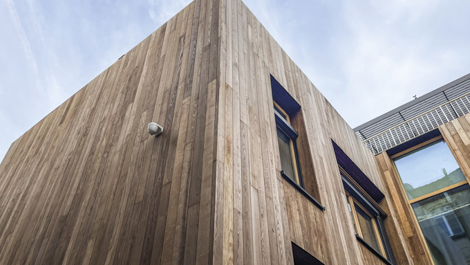 Modern building corner clad in vertical wooden panels with windows and a security camera under a partly cloudy sky.