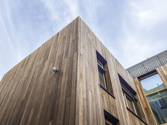 Modern building corner clad in vertical wooden panels with windows and a security camera under a partly cloudy sky.