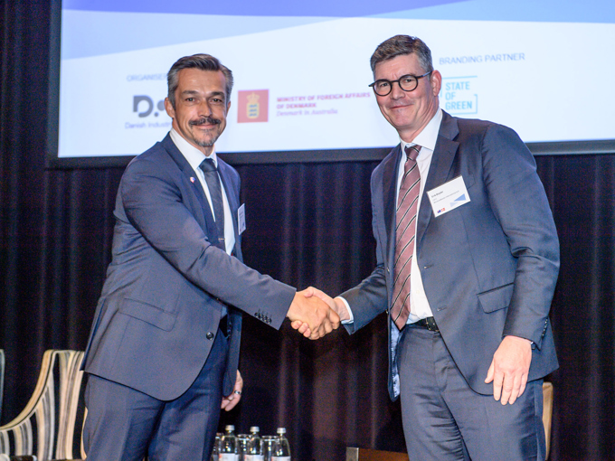 Two men in suits shaking hands at a formal event with a presentation screen and water bottles in the background.