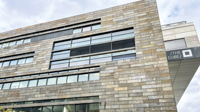 Modern building facade featuring horizontal stone panels and large windows, with "THE CUBE" signage on the side.