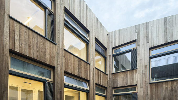 Modern wooden-clad building with large rectangular windows, some illuminated interiors, and an overcast sky above a courtyard.