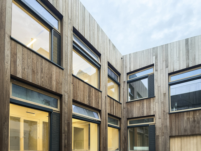 Modern wooden-clad building with large rectangular windows, some illuminated interiors, and an overcast sky above a courtyard.