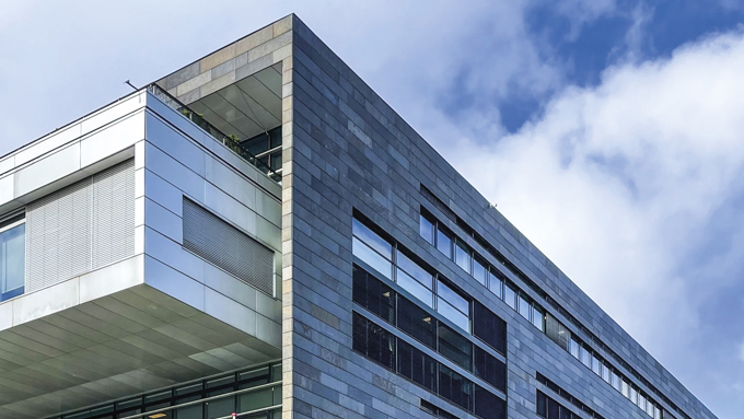 Modern building with a sleek stone facade and large glass windows, under a partly cloudy sky. Trees and cars are in the foreground.