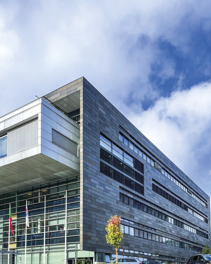 Modern building with a sleek stone facade and large glass windows, under a partly cloudy sky. Trees and cars are in the foreground.
