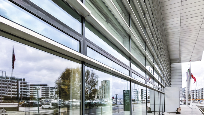 Modern building exterior featuring large glass windows and horizontal slats, reflecting surrounding urban landscape and flags.