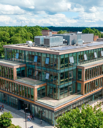 A modern, multi-story building with large glass windows, solar panels, and mechanical ventilation surrounded by trees and people walking on a sunny day.
