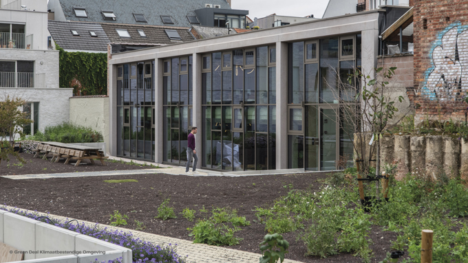 A modern building with large glass windows is set in a garden, featuring greenery and wooden seating. A person walks nearby.
