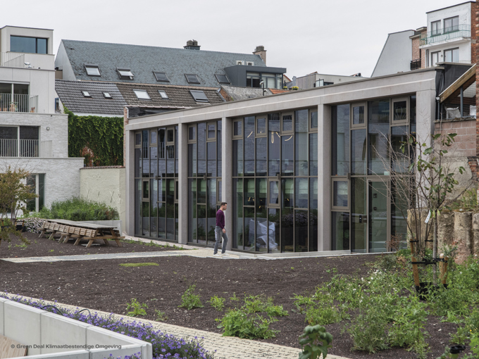 A modern building with large glass windows is set in a garden, featuring greenery and wooden seating. A person walks nearby.